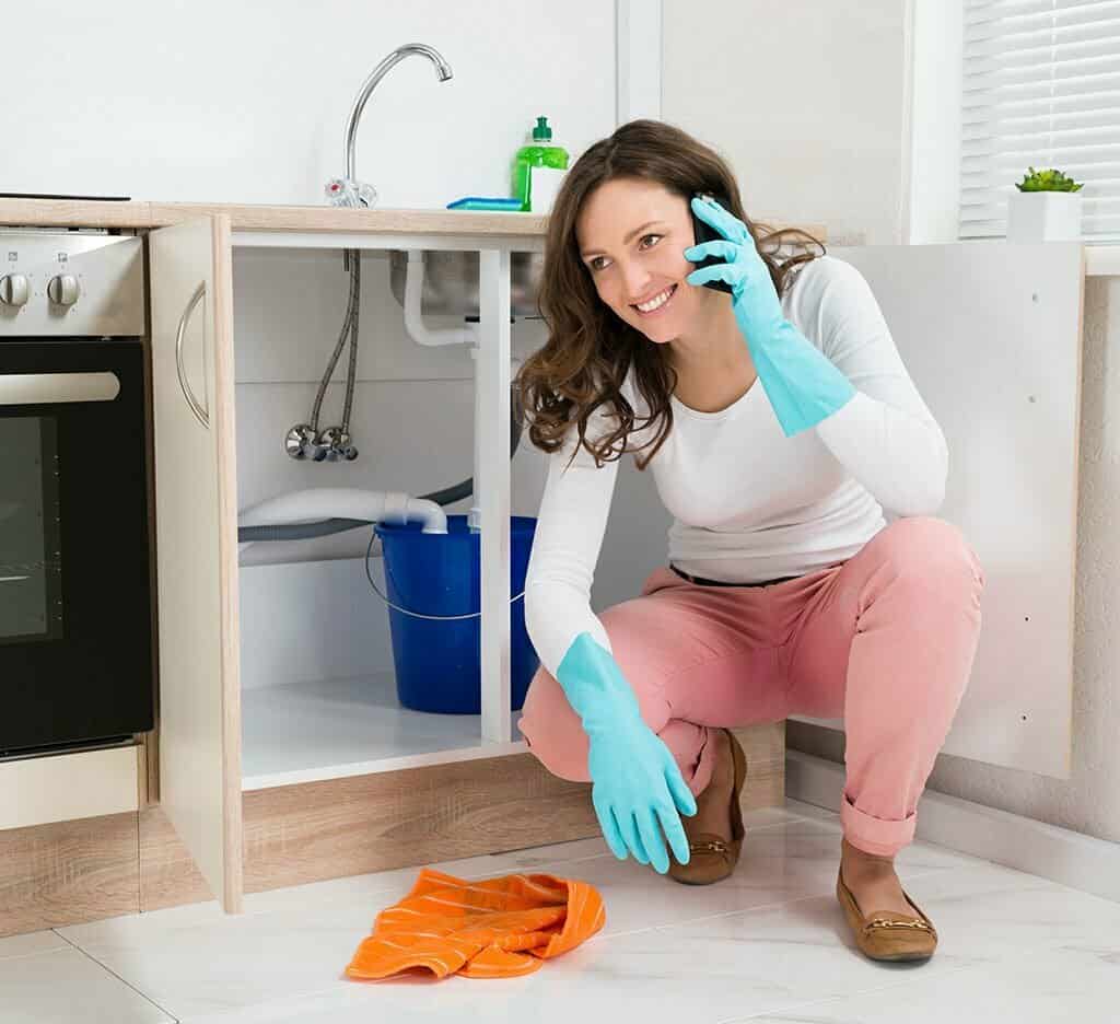 Woman crouching down next to blocked and overflowing sink with spill on the phone